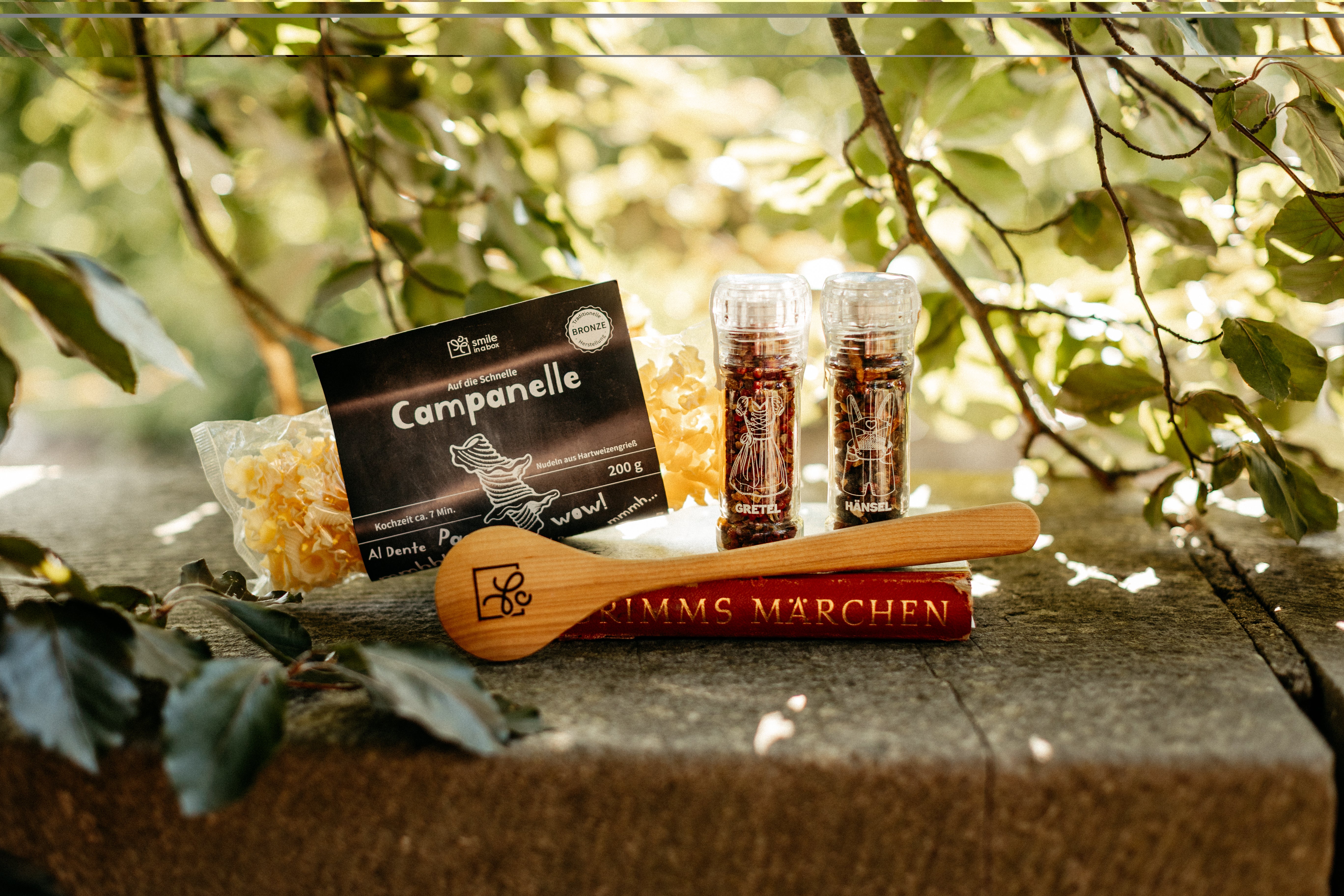 Spätsommerliches Ambiente - Salz-und-Pfeffer-Set, hölzerner Kochlöffel und Campanelle Pasta um Märchenbuch arrangiert. Eine Geschenkbox für Kochliebhaber und Märchenfans.