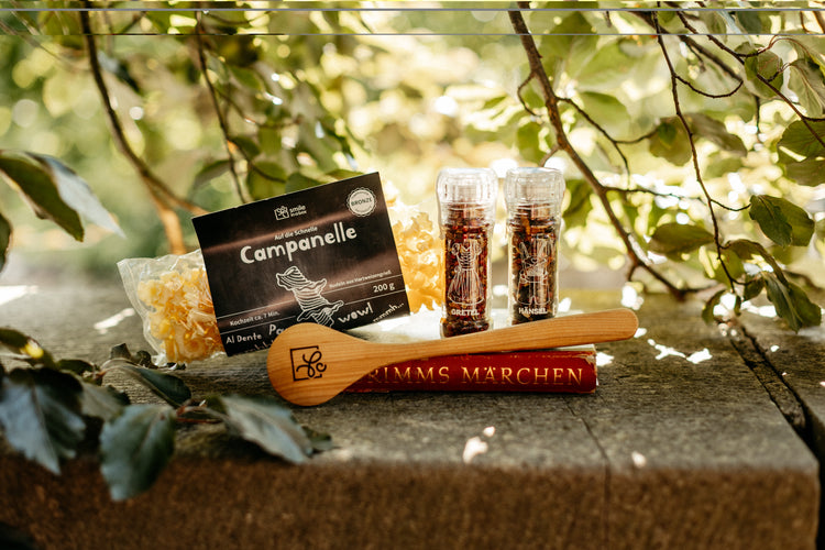 Spätsommerliches Ambiente - Salz-und-Pfeffer-Set, hölzerner Kochlöffel und Campanelle Pasta um Märchenbuch arrangiert. Eine Geschenkbox für Kochliebhaber und Märchenfans.